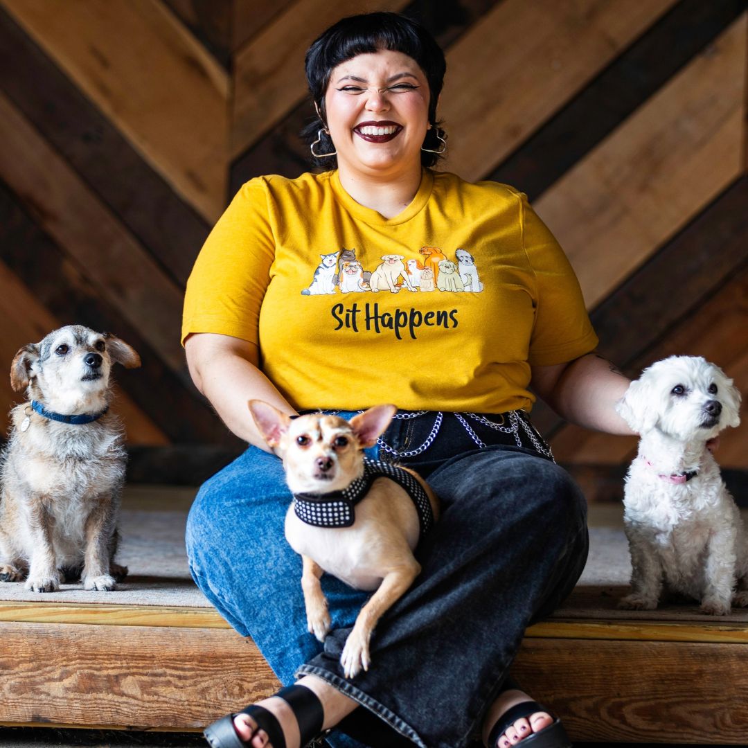 Dog Mom wearing a heather mustard shirt that says sit happens. She is with three little dogs sitting.