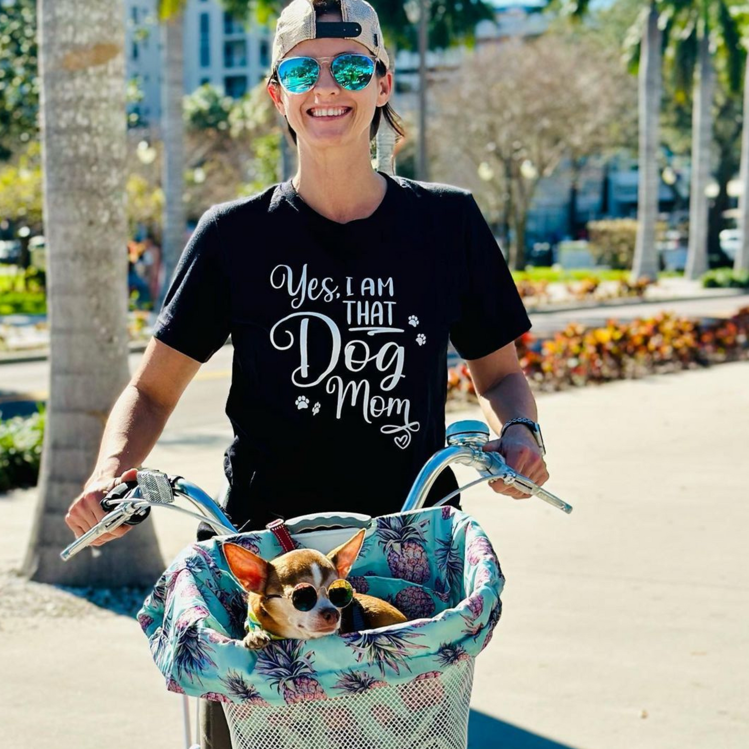 Female wearing a black shirt that says 'Yes, I Am that Dog Mom". She is riding a bike with her cute rescue dog in a basket on the front of the bike.