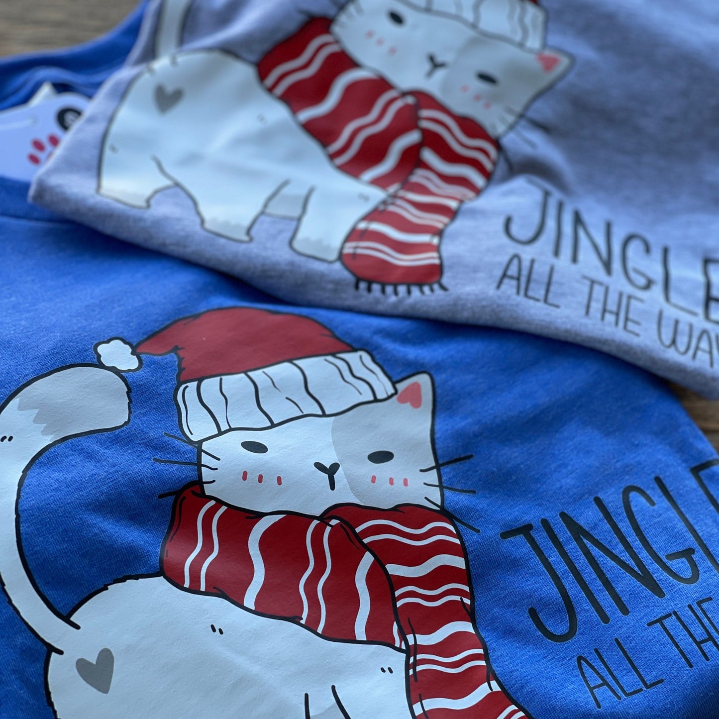 Close up picture of two shirts, one gray and one blue that features a Christmas Cat with the text Jingle All The Way