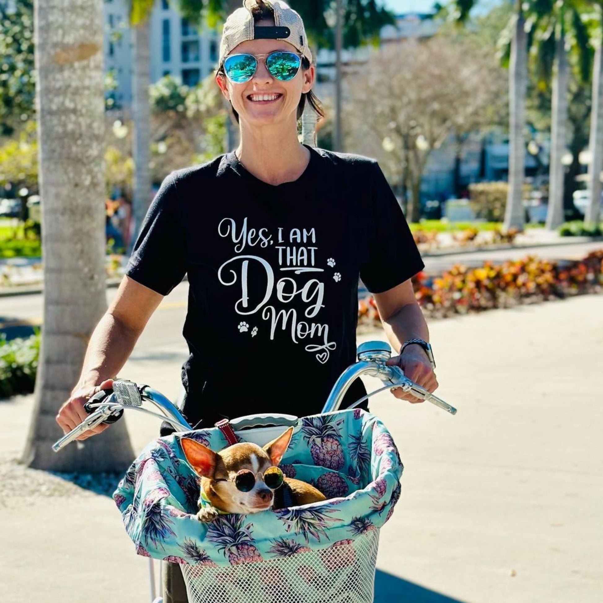 A young woman riding a bike. She is wearing a black shirt that says, "Yes, I'm that Dog Mom". She has a cool dog riding in basket in front.