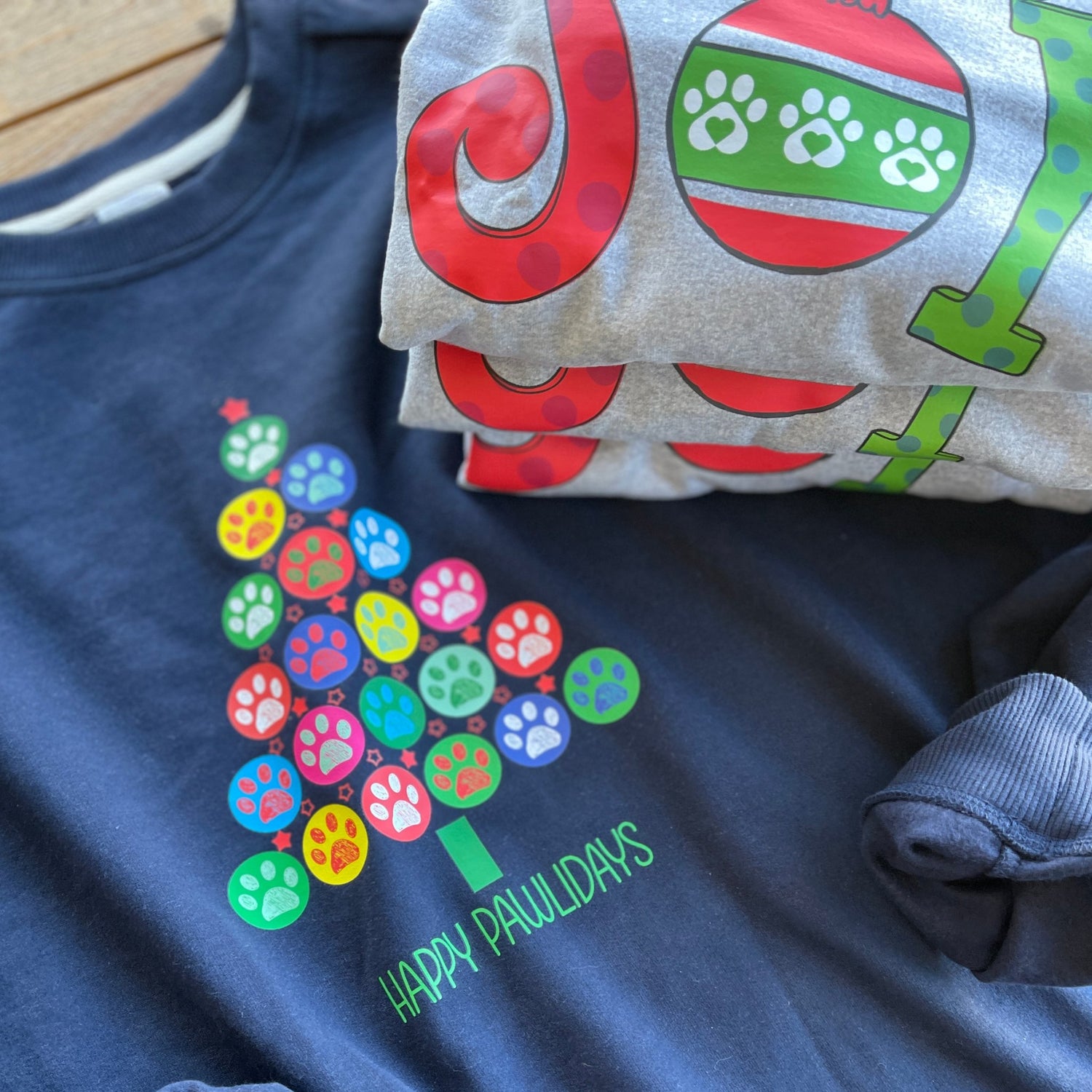 Festive Paws Holiday Collection featuring a navy blue sweatshirt with a christmas tree made of colorful dog paws and a grey sweatshirt that has the text JOY with the O as an christmas ornament with dog paws