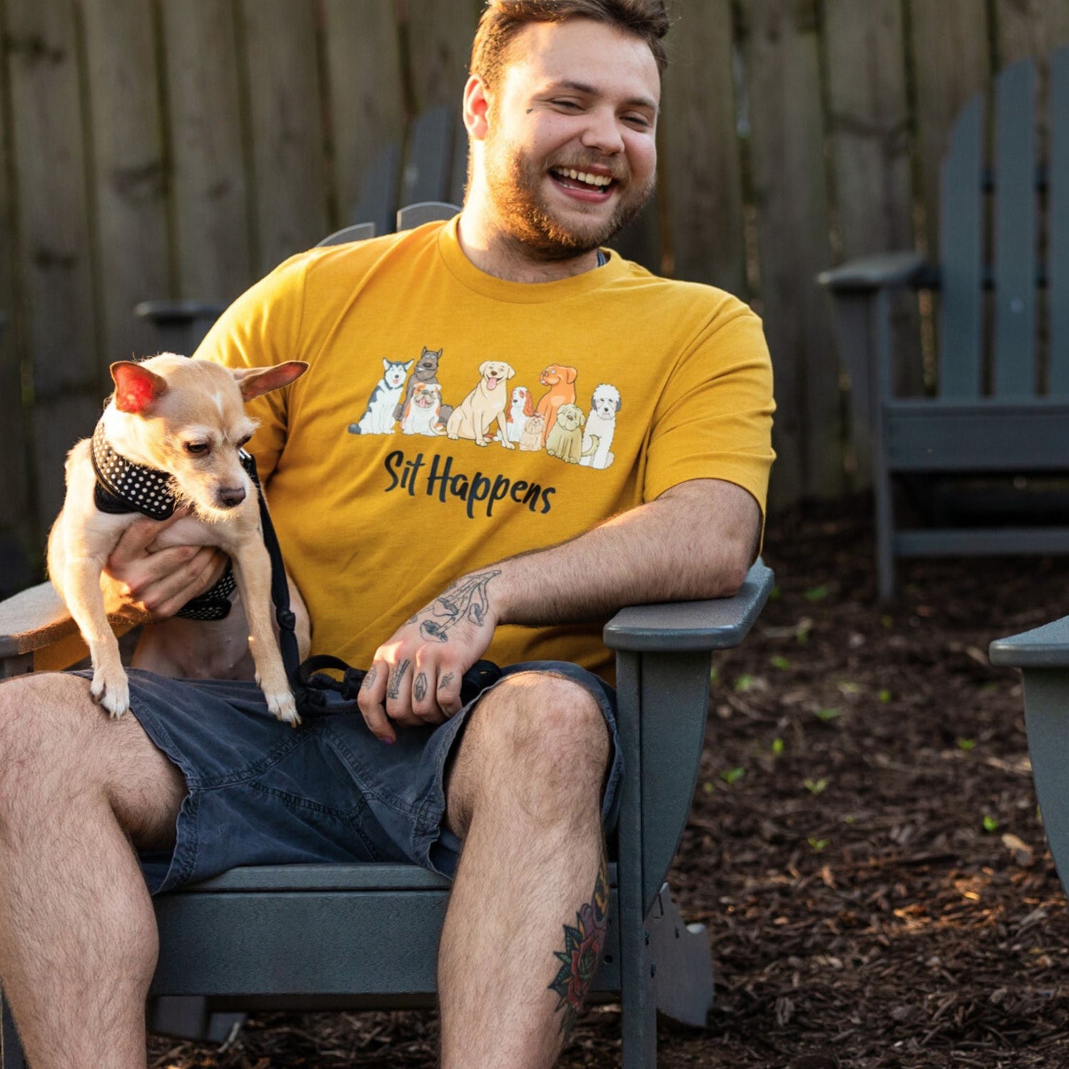 A guy sitting in an Adirondack chair with his dog. He is wearing a Heather Mustard Luv the Paw T-shirt that says "Sit Happens" with an array of dogs looking cute and sitting. 