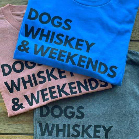 3 Shirts folded on a table one in blue, sunset and gray colors with Dogs, Whiskey & Weekends in black text.