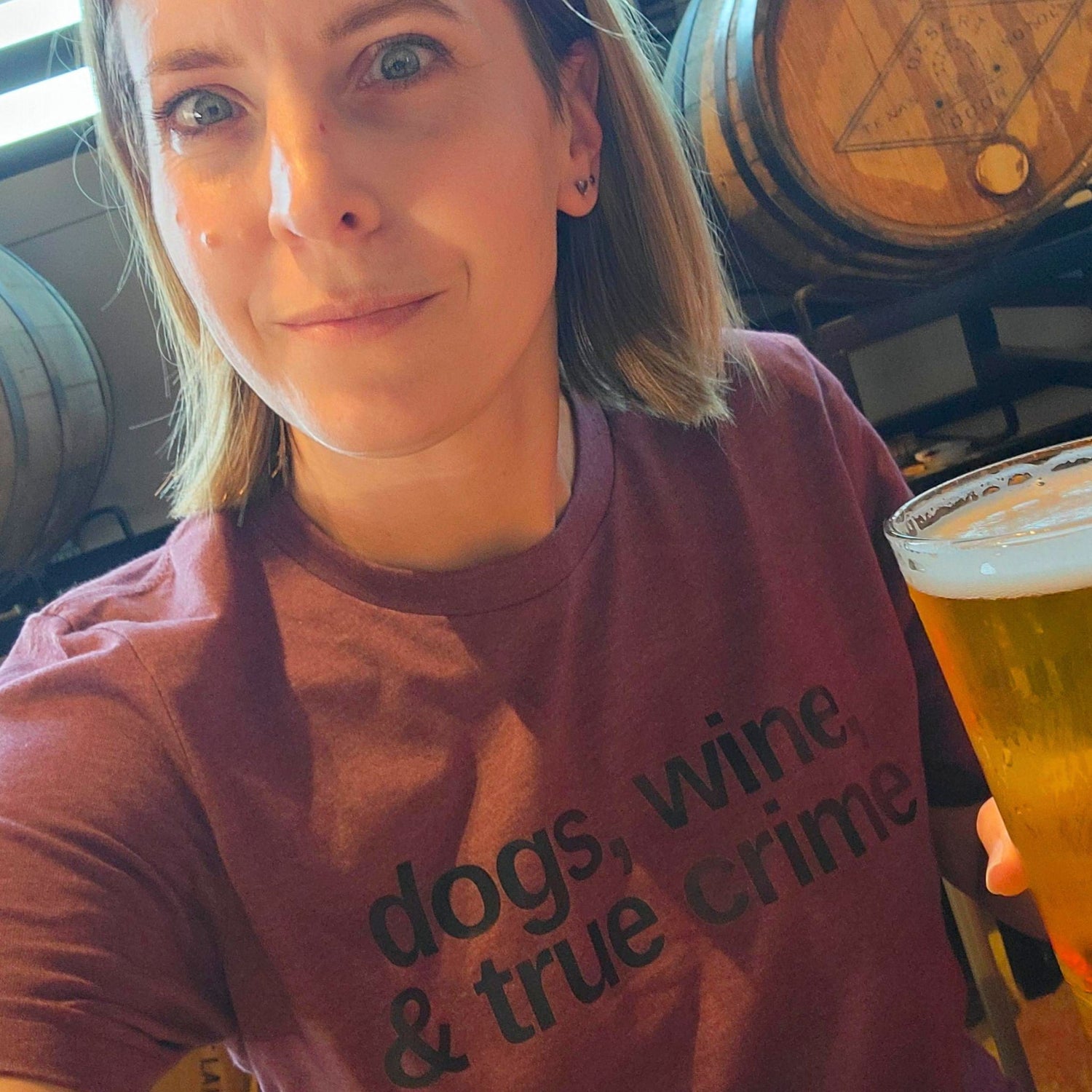 Person wearing a shirt with 'dogs, wine & true crime' text, holding a beer glass.