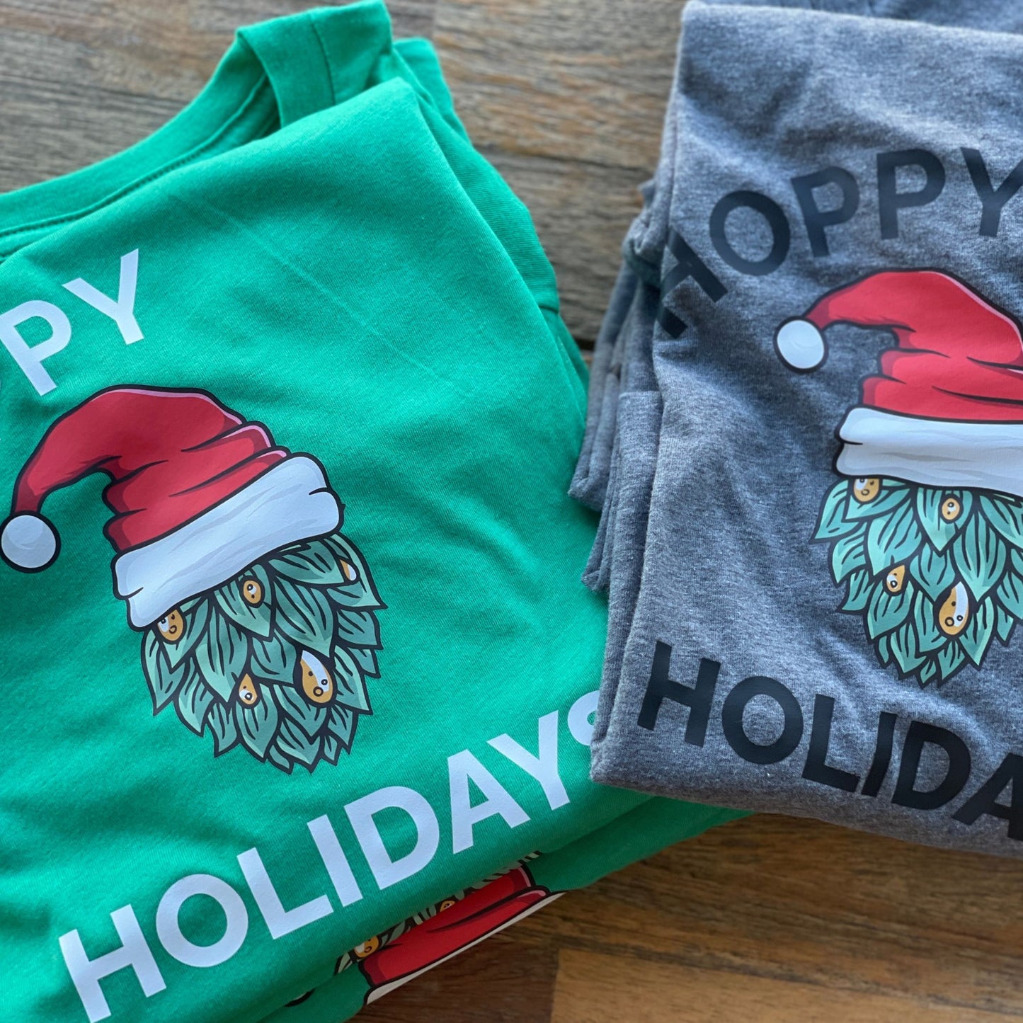 Green and Grey t-shirts folded on a table. The shirts have a hop in the middle with a santa hat with text the reads Hoppy Holidays