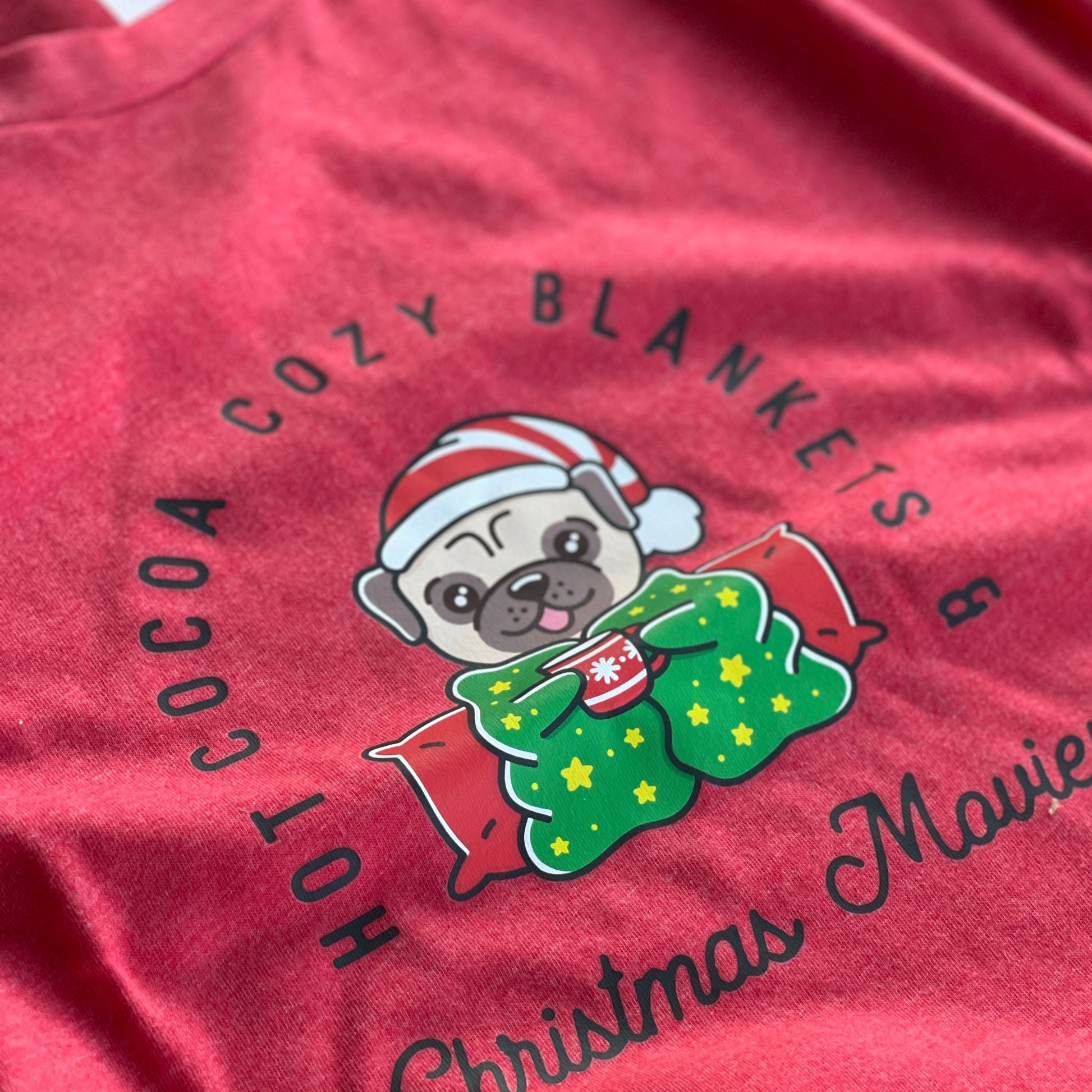 Heather red Christmas shirt featuring a pug wrapped in a blanket with a mug of hot cocoa in text around the cute dog is 'Hot Cocoa Cozy Blankets & Christmas Movies'