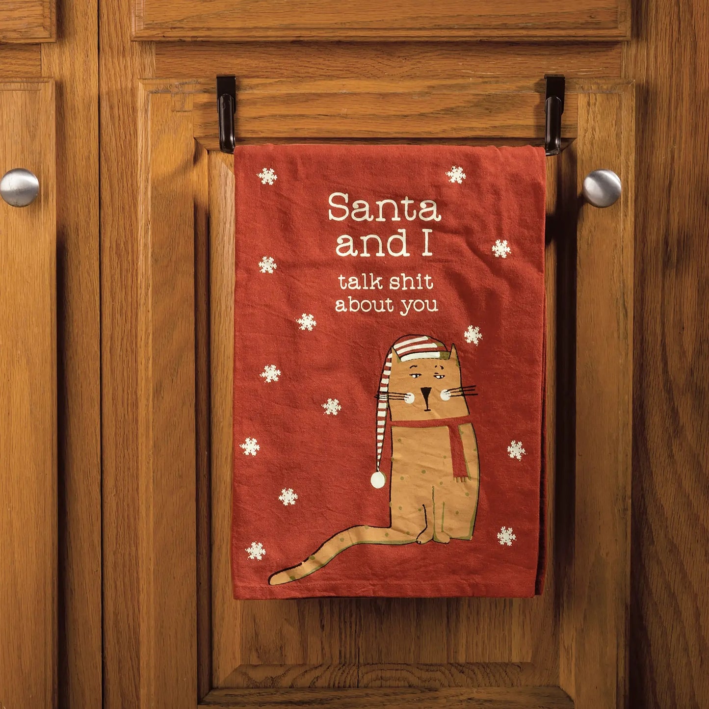 Funny red Christmas kitchen towel with cat wearing Santa hat and text “Santa and I talk shit about you” hanging on cabinet door.