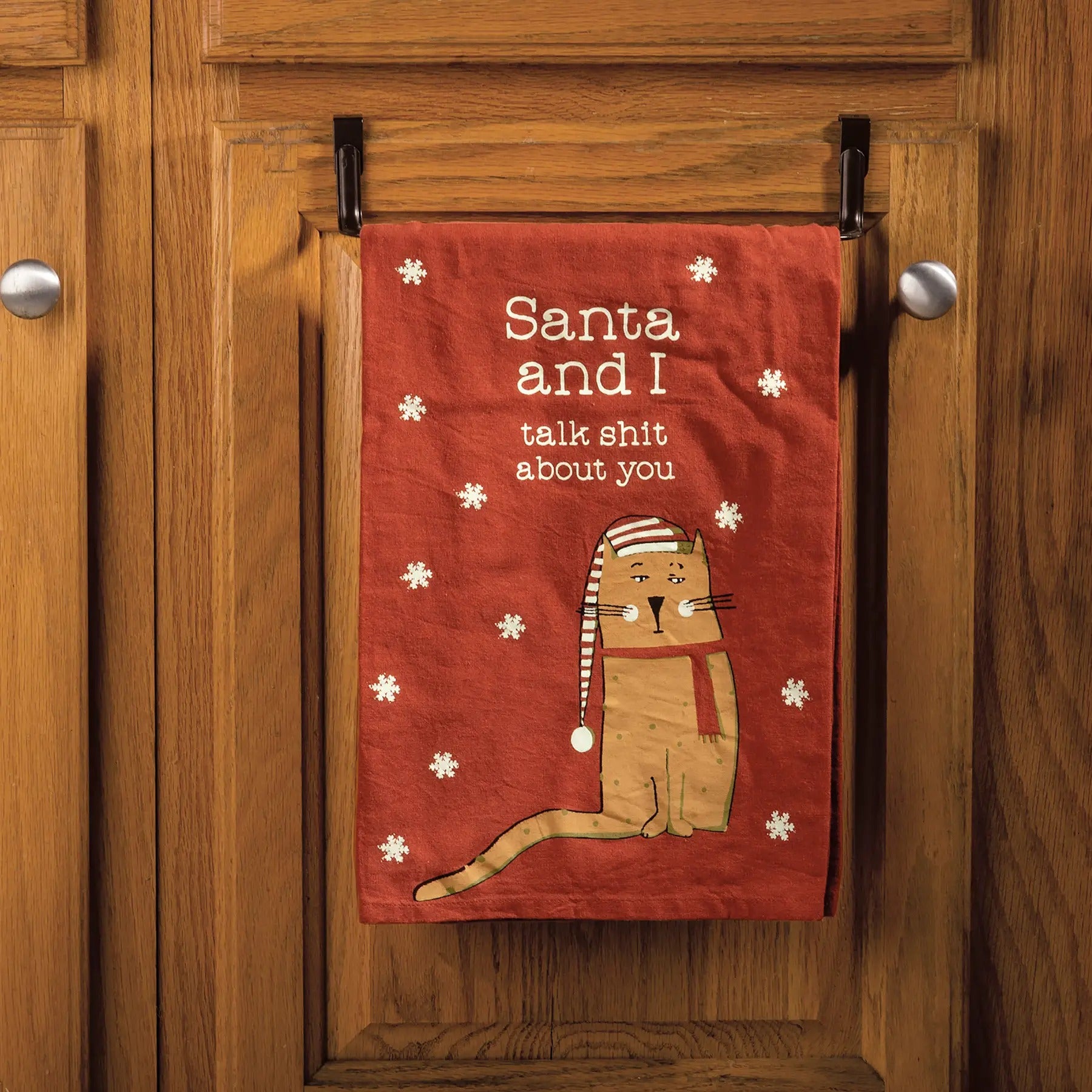 Funny red Christmas kitchen towel with cat wearing Santa hat and text “Santa and I talk shit about you” hanging on cabinet door.