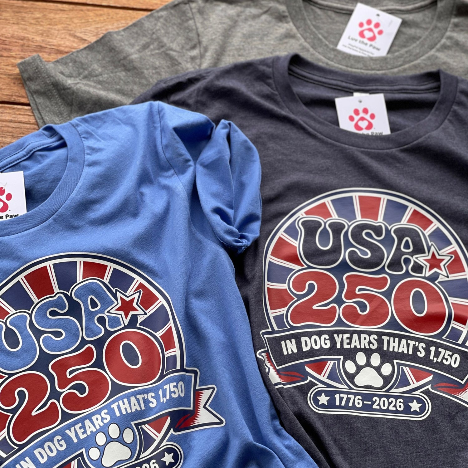 Three t-shirts with 'USA 250' design on a wooden surface