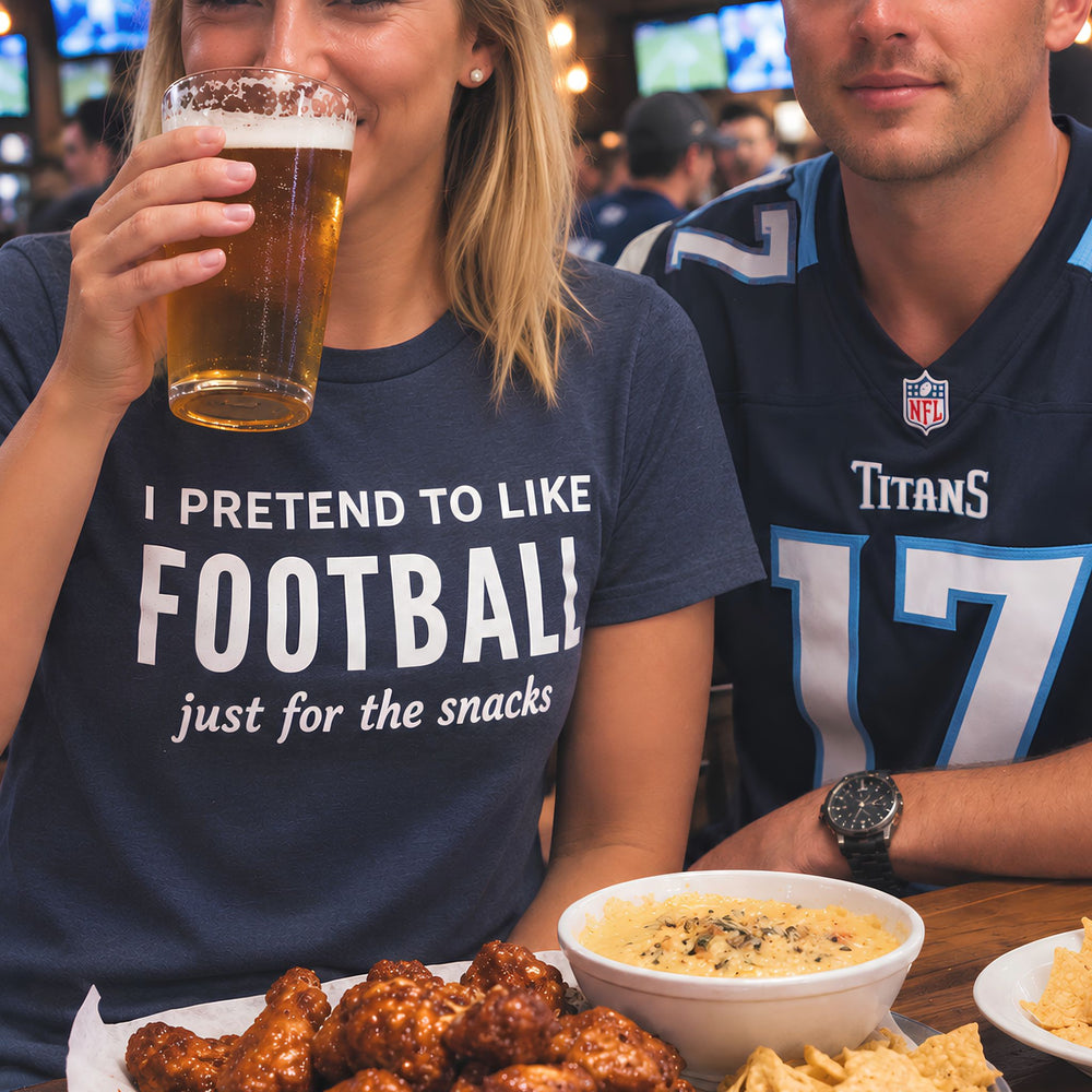 Blonde woman wearing a “I Pretend to Like Football Just for the Snacks” T-shirt, enjoying a beer with a football fan at a sports bar, surrounded by wings, chips and queso.