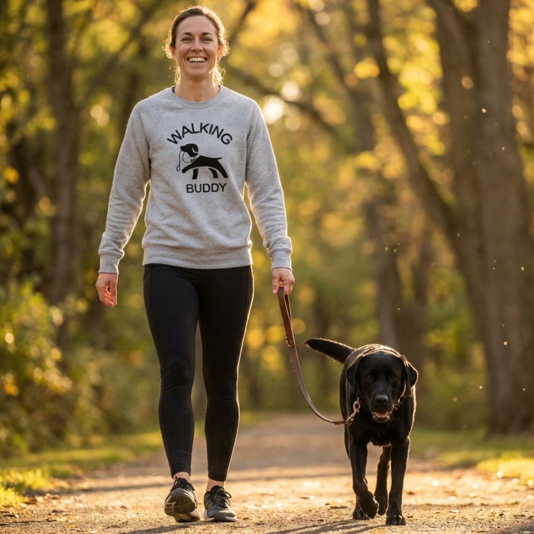 Woman walking a black dog on a path in a park with trees in the background. The woman is wearing a Snuggle Buddy Sweatshirt from Luv the Paw.