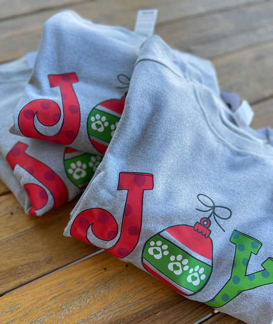 Athletic gray sweatshirts folded on a wooden table. They have 'Joy' in a whimsical text and the "O" is an ornament with paw prints. Perfect for the holiday loving pet owner.